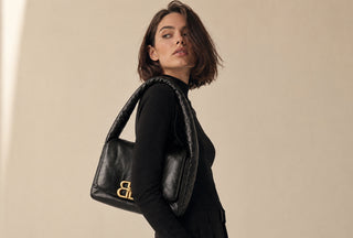 A woman in a black outfit carries a black Balenciaga leather shoulder bag with gold-tone 'BB' lettering, set against a plain beige background.