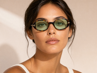 A close-up portrait of a woman wearing Tom Ford oval sunglasses with tortoiseshell frames and green-tinted lenses against a warm beige background.
